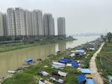 A river flows through an urban area, bordered by large residential high-rises on one side and a stretch of green land with makeshift tents or shelters on the other. The sky is overcast, casting a gray tint over the scene. A bridge is visible in the distance, and small boats are docked along the riverbank.