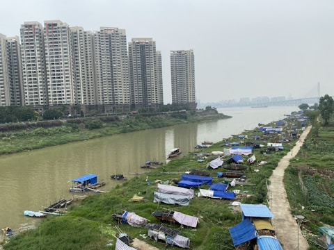 A river flows through an urban area, bordered by large residential high-rises on one side and a stretch of green land with makeshift tents or shelters on the other. The sky is overcast, casting a gray tint over the scene. A bridge is visible in the distance, and small boats are docked along the riverbank.