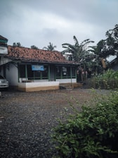 A small, single-story building with a tiled roof is surrounded by tropical plants, including banana and palm trees. The structure has a sign that reads 'Al-Fatih' and is set against an overcast sky. The foreground features a gravel lot and green foliage.