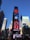 A bustling urban scene featuring a tall building adorned with multiple bright electronic billboards advertising well-known brands, surrounded by modern skyscrapers. An American flag waves prominently on a pole in the foreground.