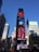A bustling urban scene featuring a tall building adorned with multiple bright electronic billboards advertising well-known brands, surrounded by modern skyscrapers. An American flag waves prominently on a pole in the foreground.
