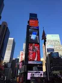 A bustling urban scene featuring a tall building adorned with multiple bright electronic billboards advertising well-known brands, surrounded by modern skyscrapers. An American flag waves prominently on a pole in the foreground.