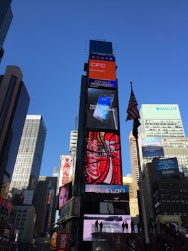 A bustling urban scene featuring a tall building adorned with multiple bright electronic billboards advertising well-known brands, surrounded by modern skyscrapers. An American flag waves prominently on a pole in the foreground.