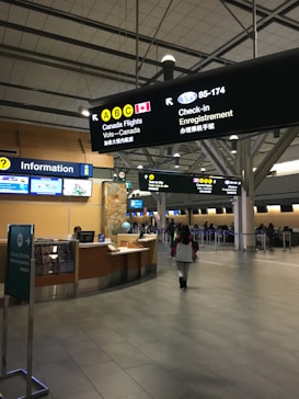 The interior of an airport terminal includes signage for Canadian flights, check-in counters, and an information desk. A person is walking towards the check-in area, and there are several illuminated signs providing directions. The setting is clean and modern with high ceilings.