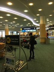 A person stands near a luggage cart in an airport, with signs indicating information desks nearby. Several people are sitting on the floor, some using their phones. The ceiling has modern lighting fixtures, and there is a sign for currency exchange in the background.