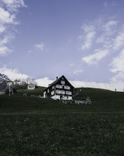 A rustic, dark-colored chalet-style house surrounded by lush green grass on a hillside. The sky is clear with scattered soft clouds, and there are faint outlines of distant mountains in the background. A small wooden building is nearby, and there is an outdoor sitting area with a fence around the property.