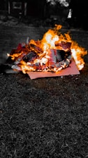 A vibrant fire burns intensely on pieces of wood or debris, with flames leaping high and producing bright orange and yellow hues. The background is mostly dark, highlighting the vivid flames and charred material.