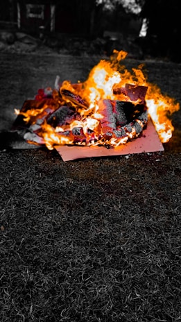 A vibrant fire burns intensely on pieces of wood or debris, with flames leaping high and producing bright orange and yellow hues. The background is mostly dark, highlighting the vivid flames and charred material.