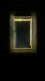 A rectangular mirror with a distressed golden-brown frame, reflecting a dim light. The frame has a worn and tarnished appearance, giving it an antique or vintage feel, while the surrounding darkness enhances its luminous quality.