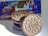 A stack of chocolate sandwich cookies with cream filling is prominently displayed, with a focus on the intricate logo and pattern on the top cookie. In the background, there is a partially visible blue and red package with an image of the cookies and nutritional information.