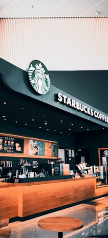 A modern Starbucks coffee shop with a black and wood-themed counter. The menu is prominently displayed above the counter, and various coffee-making machines and accessories are organized neatly. The store features bright lighting, with a minimalist design style, and the iconic Starbucks logo is visible.