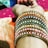 A close-up of a wrist adorned with multiple colorful bangles, featuring intricate gold detailing and beads. The hand is resting against a backdrop of vibrant, patterned fabrics in orange and pink.
