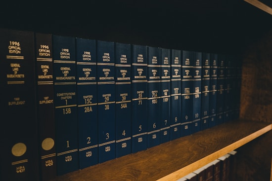 A row of dark blue hardcover books arranged on a wooden shelf, with uniform lettering on their spines indicating they are volumes of the General Laws of Massachusetts. The books appear to be reference materials organized in a comprehensive collection.