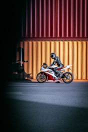 A motorcyclist, dressed in black protective gear, is sitting on a sporty Yamaha motorcycle. The motorcycle has a red and white color scheme and is parked on a wide, empty road in front of large, brightly colored shipping containers in yellow and red hues.
