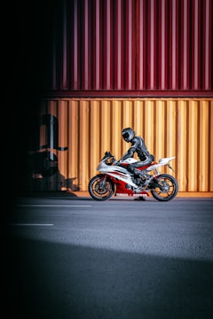 A motorcyclist, dressed in black protective gear, is sitting on a sporty Yamaha motorcycle. The motorcycle has a red and white color scheme and is parked on a wide, empty road in front of large, brightly colored shipping containers in yellow and red hues.