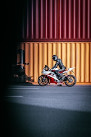 A motorcyclist, dressed in black protective gear, is sitting on a sporty Yamaha motorcycle. The motorcycle has a red and white color scheme and is parked on a wide, empty road in front of large, brightly colored shipping containers in yellow and red hues.