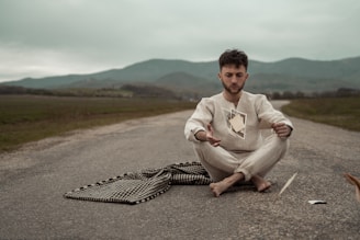 A person sits cross-legged on an empty road, appearing contemplative. Floating in front of them are several photographs. The scene is set against a backdrop of rolling hills under a cloudy sky, creating an atmospheric and introspective tone.