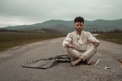 A person sits cross-legged on an empty road, appearing contemplative. Floating in front of them are several photographs. The scene is set against a backdrop of rolling hills under a cloudy sky, creating an atmospheric and introspective tone.
