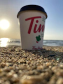 Close-up of coffee pouring into a clear glass cup with a sandy beach and gentle waves in the distance.
