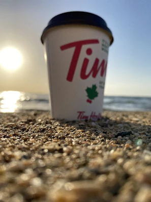 Close-up of coffee pouring into a clear glass cup with a sandy beach and gentle waves in the distance.