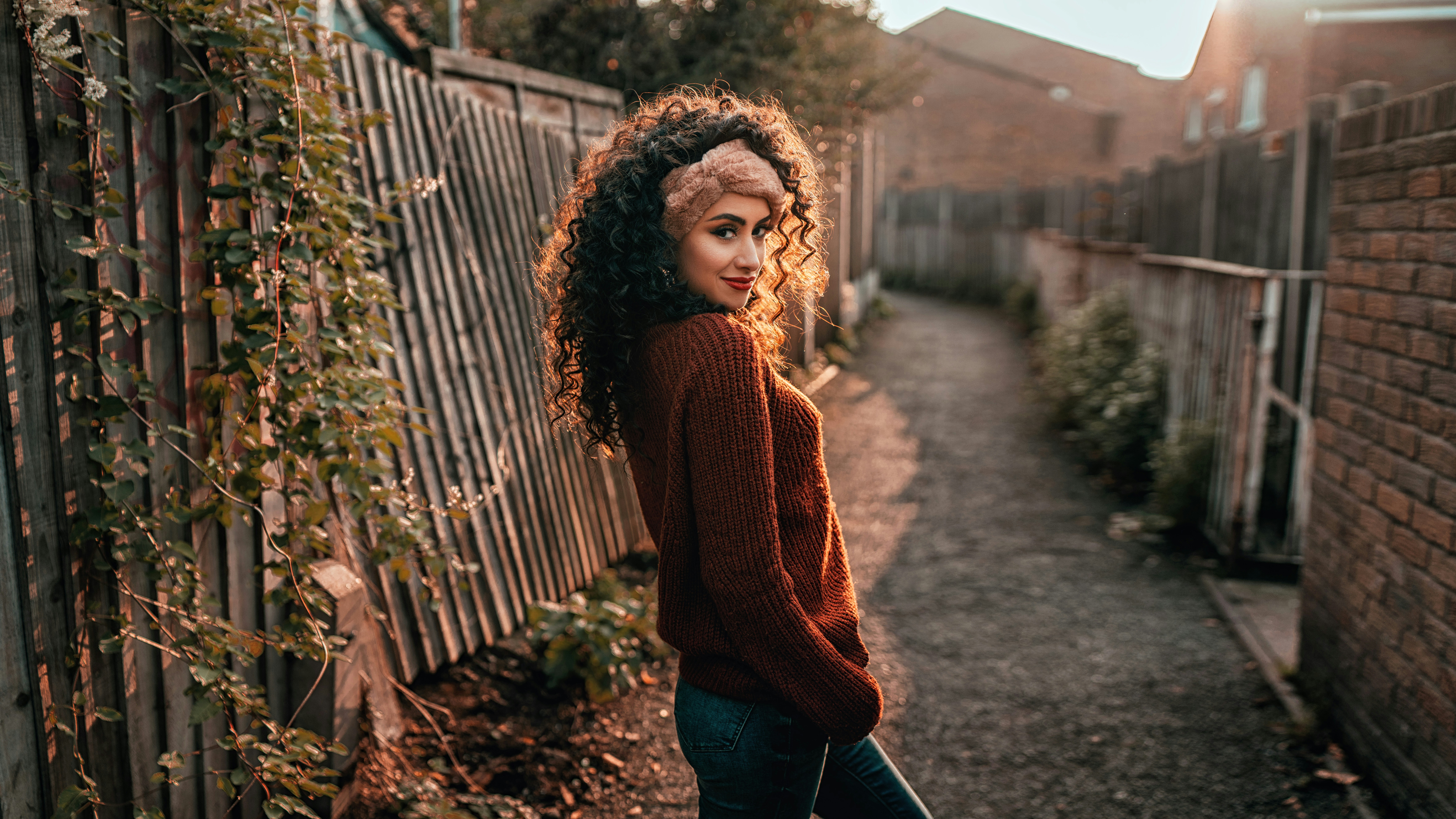 woman in brown knit sweater and blue denim jeans standing near brown wooden fence during daytime