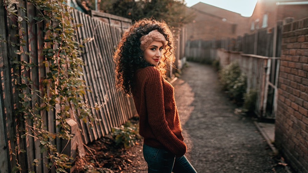 woman in brown knit sweater and blue denim jeans standing near brown wooden fence during daytime