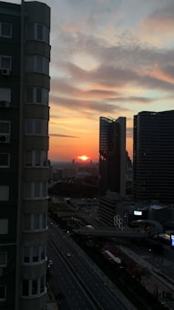 A cityscape at sunset with buildings and a vibrant sky that includes shades of orange, pink, and blue. Tall modern skyscrapers frame the sun as it sets on the horizon, casting shadows over the urban landscape. The scene is observed from a high vantage point, capturing the intricate details of the architecture and streets below.