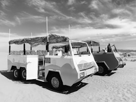 Two off-road vehicles with canopies are parked on a sandy landscape under a partly cloudy sky. The vehicles have a rugged design with multiple wheels and open sides. Some mid-sized mountains or hills are visible in the background.