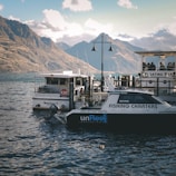 A serene lakeside scene with several boats docked at a marina, part of a picturesque landscape featuring calm waters and mountains in the background. A boat labeled 'Fishing Charters' and another offering a '90 Minute Lake Cruise' are visible, alongside a floating bar. There is a sense of leisure with people aboard the bar, and the distant mountains under a partly cloudy sky add to the peaceful atmosphere.
