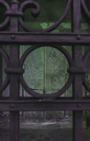 A view through an ornate metal gate, highlighting a wall covered with numerous plaques containing names and inscriptions. The gate's design features circular patterns, and the background exudes a somber atmosphere with aged and worn textures on the wall.