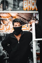 A young person stands in front of a poster with bold lettering and vivid imagery, wearing a black jacket and a face covering. The background features a dramatic design with a horror theme, emphasizing vivid artwork and text.