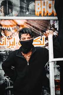 A young person stands in front of a poster with bold lettering and vivid imagery, wearing a black jacket and a face covering. The background features a dramatic design with a horror theme, emphasizing vivid artwork and text.