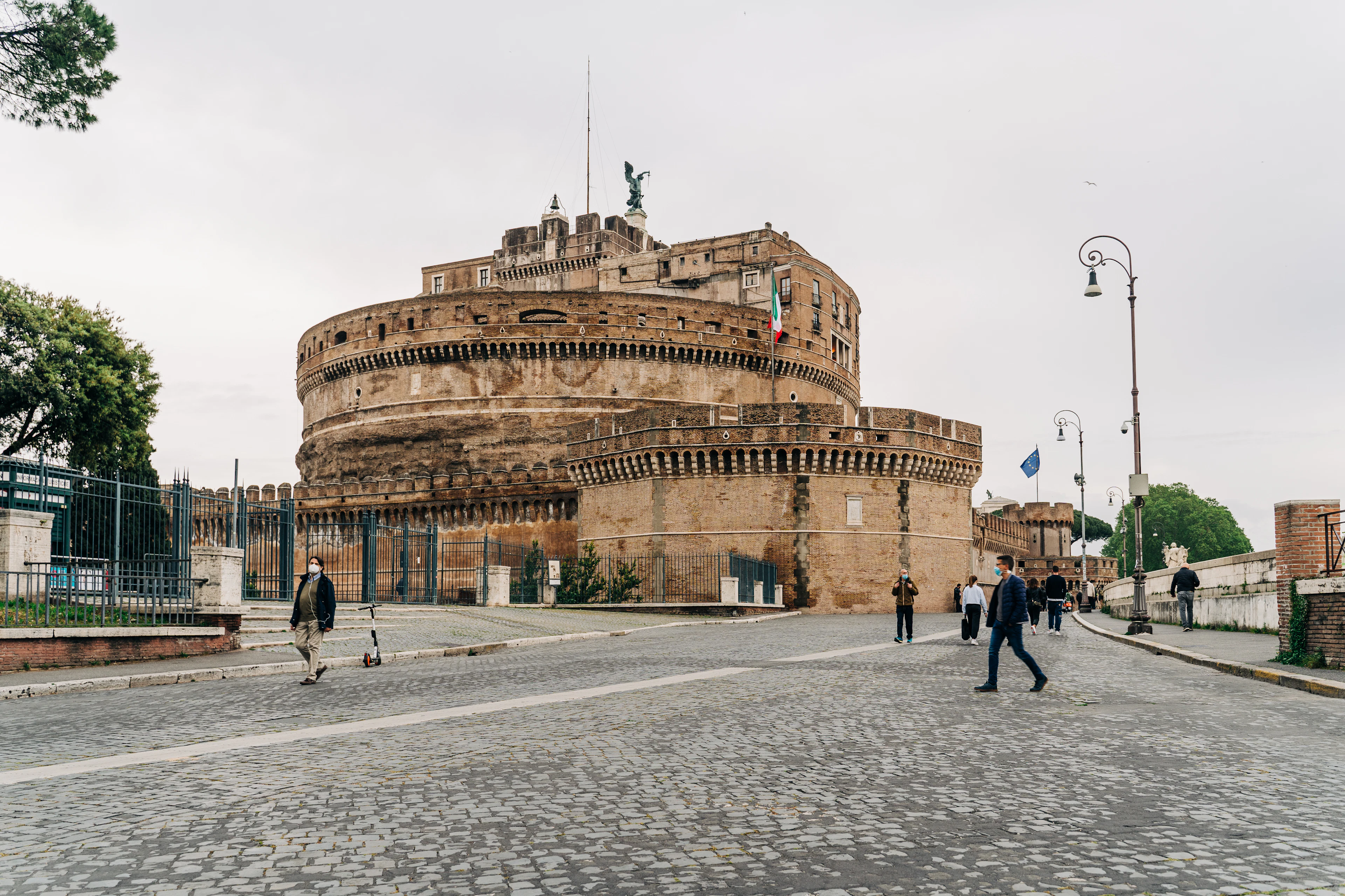 The circular brick drum of Castel Sant'Angelo rising above the cobbled riverside road, with people crossing in front, Rome