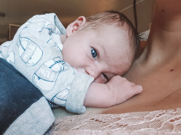 A baby rests its head on an adult's chest, wearing a light-colored onesie with a milk carton pattern. The baby's blue eyes look directly at the camera. The adult is wearing a lace pink garment.
