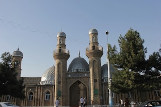 A mosque with two prominent minarets and a central dome, constructed from brick and featuring intricate patterns. The building is set against a clear blue sky with trees in the foreground and people walking nearby.