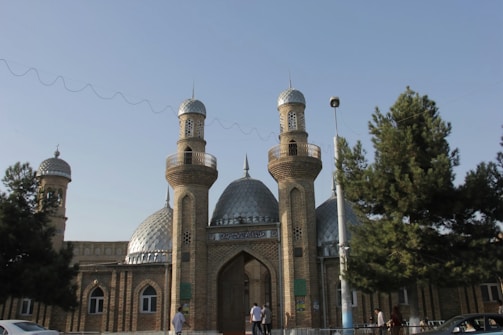 A mosque with two prominent minarets and a central dome, constructed from brick and featuring intricate patterns. The building is set against a clear blue sky with trees in the foreground and people walking nearby.