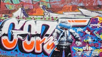 Colorful graffiti artwork on a brick wall features large, vibrant letters and a character wearing a hooded jacket. The character is holding a dripping paint roller and a brush, and the background contains abstract shapes and bright patterns. Words and phrases like 'years RIP' are also incorporated into the design.