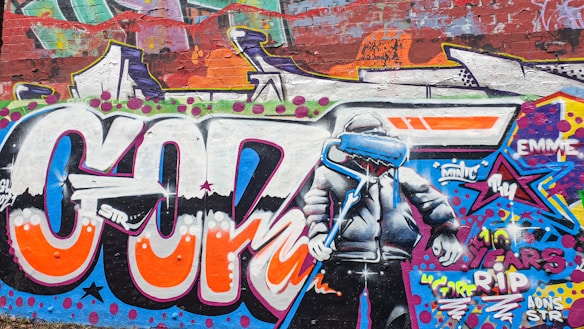 Colorful graffiti artwork on a brick wall features large, vibrant letters and a character wearing a hooded jacket. The character is holding a dripping paint roller and a brush, and the background contains abstract shapes and bright patterns. Words and phrases like 'years RIP' are also incorporated into the design.