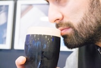 A person with a beard closely observes or smells a glass of dark beer, topped with a creamy head. The background is slightly out of focus with blurred white and blue elements, suggesting a casual indoor setting such as a bar or caf&eacute;.