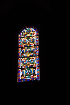 A beautifully crafted stained glass window depicting intricate patterns with vibrant colors. The design includes symmetrical floral and geometric motifs, dominated by rich reds, blues, and yellows, set against a dark background.
