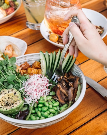 A hand holding a fork is poised over a colorful salad bowl containing sliced avocado, radish, fresh greens, edamame, cucumber slices, and mushrooms. The salad is garnished with black and white sesame seeds. In the background, there are iced beverages in glasses, including a drink with an orange tint and another with lime. The setting is a wooden table, suggesting an outdoor or casual dining environment.