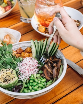 A hand holding a fork is poised over a colorful salad bowl containing sliced avocado, radish, fresh greens, edamame, cucumber slices, and mushrooms. The salad is garnished with black and white sesame seeds. In the background, there are iced beverages in glasses, including a drink with an orange tint and another with lime. The setting is a wooden table, suggesting an outdoor or casual dining environment.