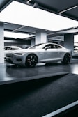 A sleek, silver sports car is parked inside a modern, well-lit showroom. The car features prominent aerodynamic lines, large black alloy wheels, and a distinctive front grille. The lighting in the room highlights the car’s glossy finish, and the minimalist design of the surroundings complements the high-tech appearance of the vehicle.