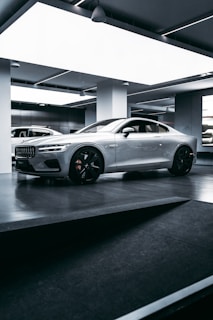 A sleek, silver sports car is parked inside a modern, well-lit showroom. The car features prominent aerodynamic lines, large black alloy wheels, and a distinctive front grille. The lighting in the room highlights the car’s glossy finish, and the minimalist design of the surroundings complements the high-tech appearance of the vehicle.
