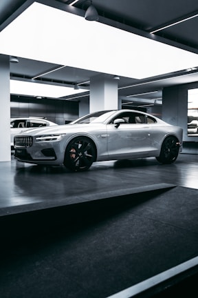 A sleek, silver sports car is parked inside a modern, well-lit showroom. The car features prominent aerodynamic lines, large black alloy wheels, and a distinctive front grille. The lighting in the room highlights the car’s glossy finish, and the minimalist design of the surroundings complements the high-tech appearance of the vehicle.