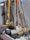 A collection of cleaning tools, including mops and brooms, is clustered together. The handles are made of wood and metal, with some featuring colored grips in yellow, orange, and blue. The mop heads are visibly made of fabric strands and the brooms have stiff bristles.