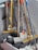 A collection of cleaning tools, including mops and brooms, is clustered together. The handles are made of wood and metal, with some featuring colored grips in yellow, orange, and blue. The mop heads are visibly made of fabric strands and the brooms have stiff bristles.
