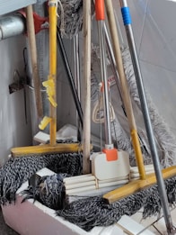 A collection of cleaning tools, including mops and brooms, is clustered together. The handles are made of wood and metal, with some featuring colored grips in yellow, orange, and blue. The mop heads are visibly made of fabric strands and the brooms have stiff bristles.