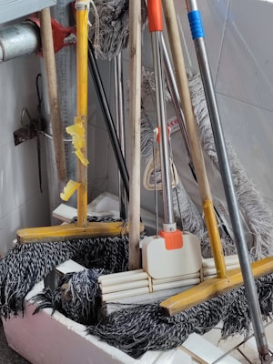 A collection of cleaning tools, including mops and brooms, is clustered together. The handles are made of wood and metal, with some featuring colored grips in yellow, orange, and blue. The mop heads are visibly made of fabric strands and the brooms have stiff bristles.