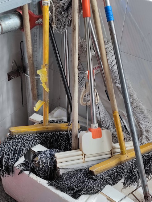A collection of cleaning tools, including mops and brooms, is clustered together. The handles are made of wood and metal, with some featuring colored grips in yellow, orange, and blue. The mop heads are visibly made of fabric strands and the brooms have stiff bristles.